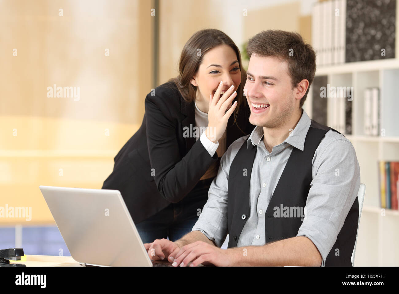 Gossip businesswoman telling secrets to the ear of a businessman sitting on a desktop at office Stock Photo
