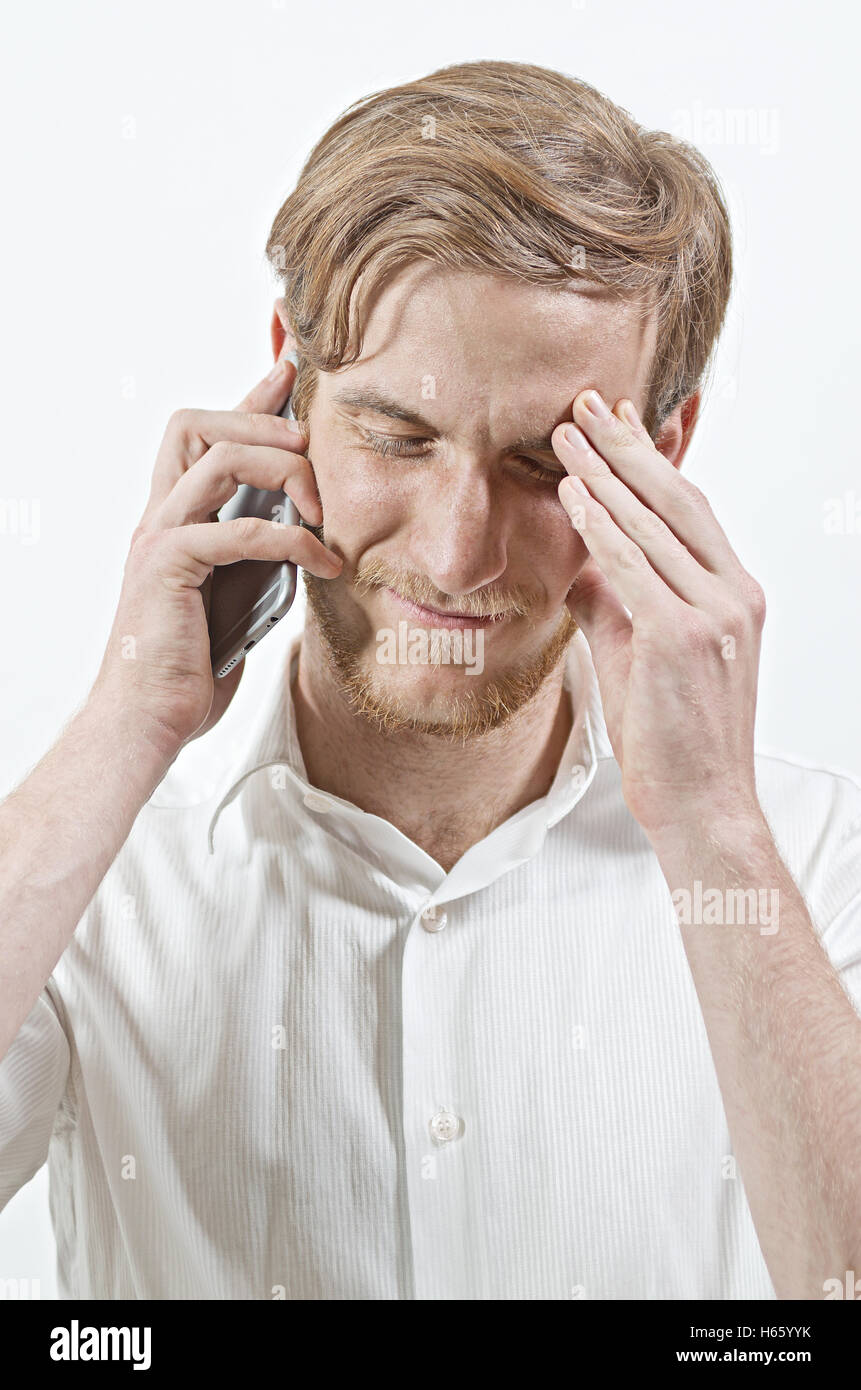 Young Adult Man in White Shirt Listening to His Phone, Touching His Head with a Hand, Receiving Bad News Stock Photo
