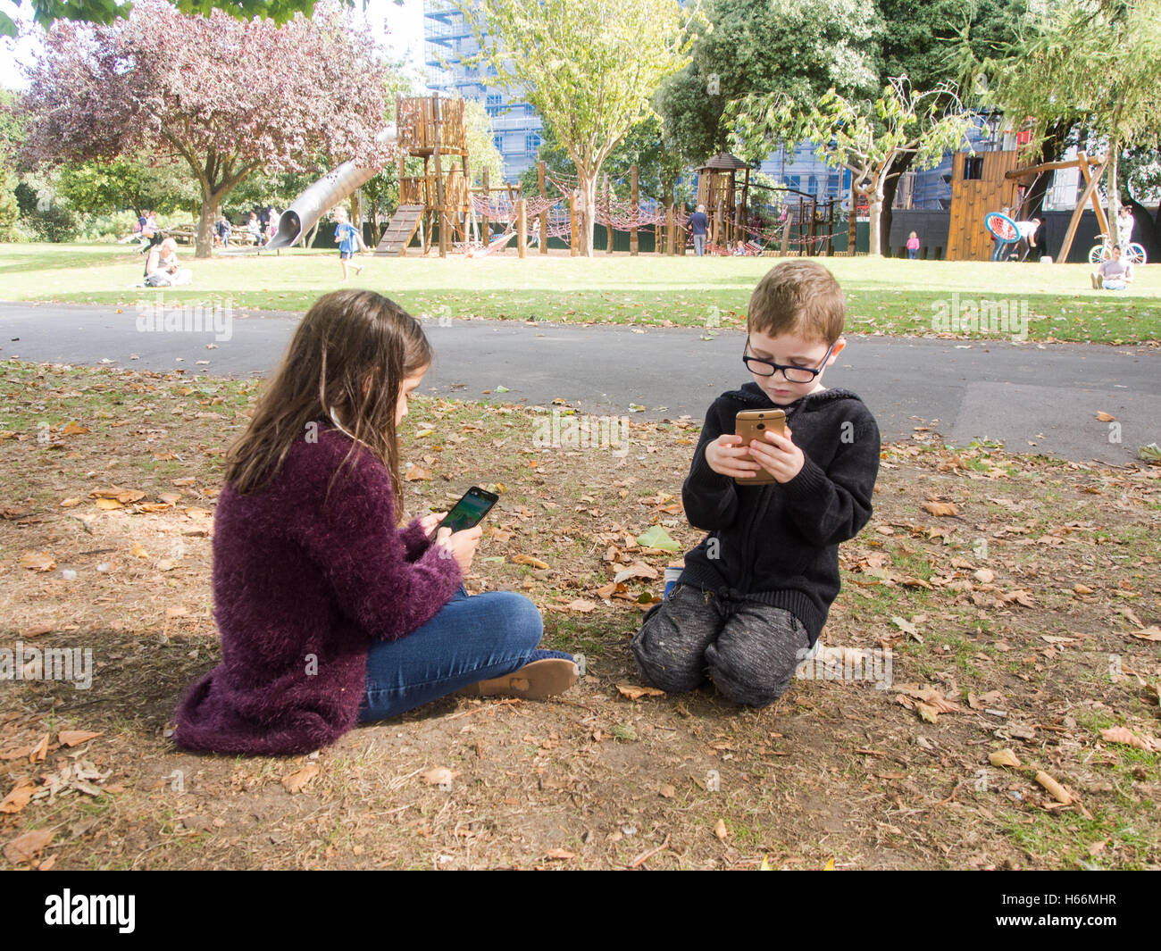 two children play games on mobile phones in a public park with a play area behind them. Stock Photo