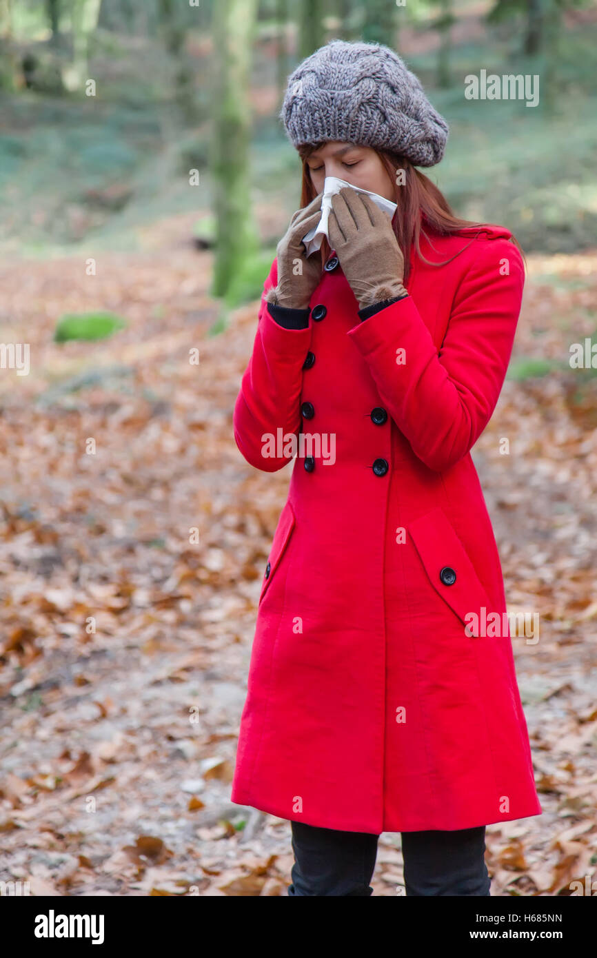 Woman with a cold or flu sneezing to a paper handkerchief on a forest wearing a red overcoat, a beanie and gloves during winter Stock Photo