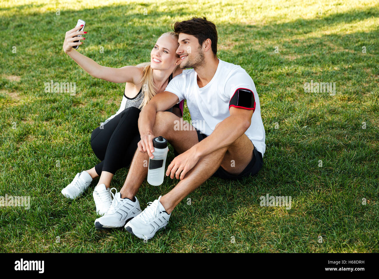 Sport man and woman making selfie in park while sitting on green grass and resting after jogging Stock Photo