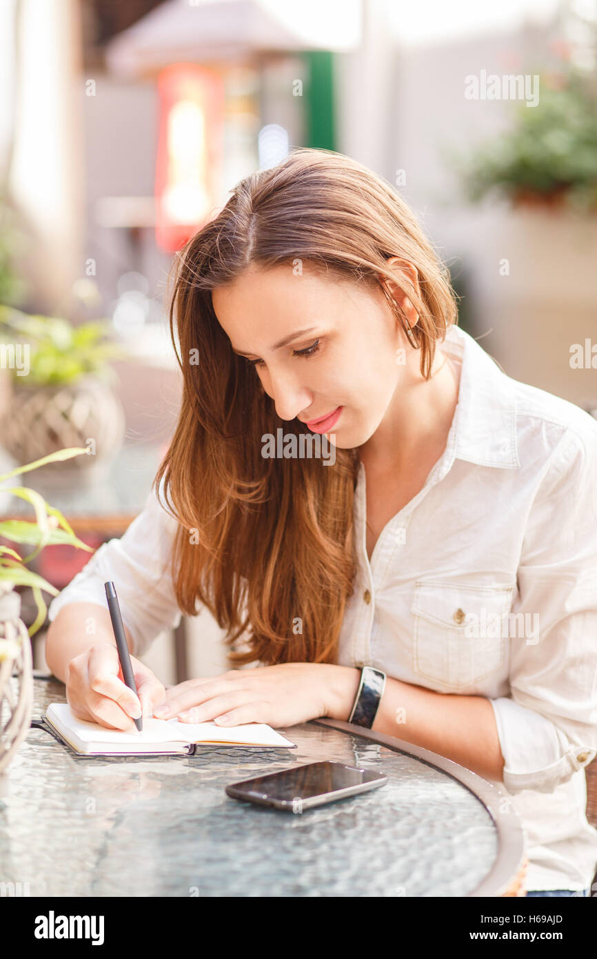 Young woman writing down into her notebook sitting at the table in restaurant Stock Photo