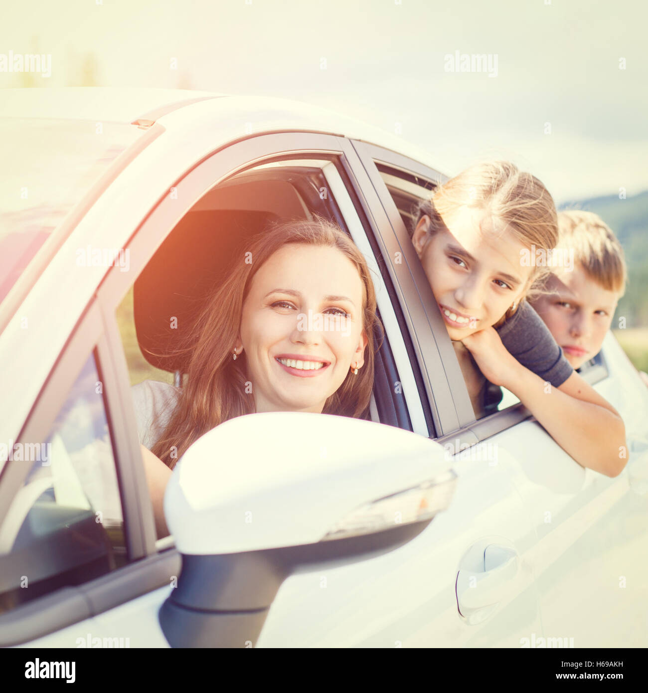 Happy young woman and her children sitting in a car and look out from windows. Family travel warm color toned image Stock Photo