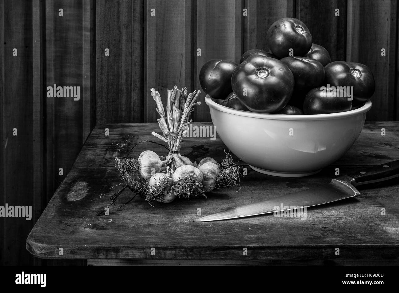 Still life in black and white  with tomatoes on white bowl with a bunch of garlic and a knife on a rustic wooden table. Stock Photo