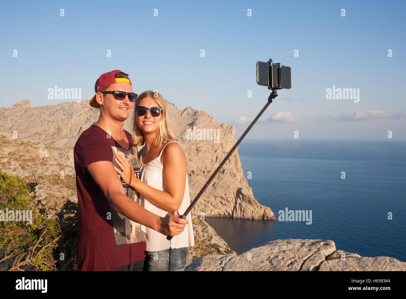One male and a female using a selfie stick and mobile phone to photograph themselves at Formentor, Mallorca, Summer 2016. Blue sky and sea behind them. Stock Photo