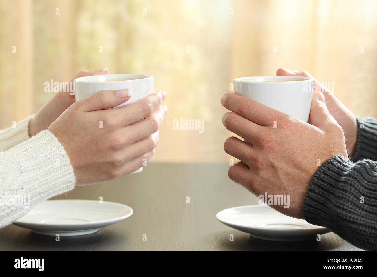 Profile of hands of a couple holding coffee cups over a table in winter in an apartment with a window in the background Stock Photo