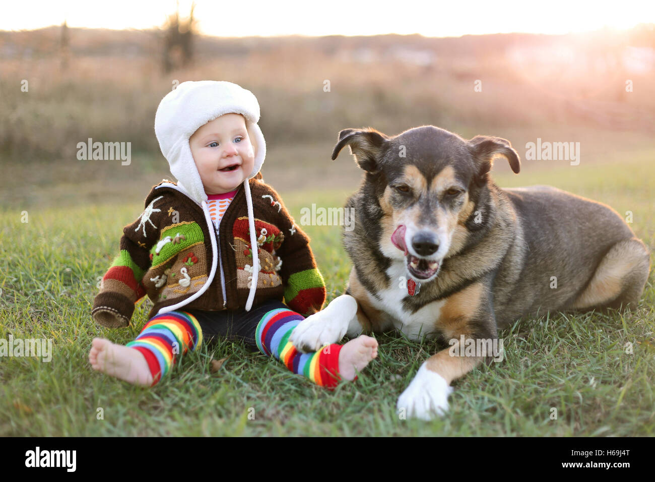 An adorable 8 month old baby girl is bundled up in a sweater and wearing a winter earflap hat looking lovinlgy at her pet German Stock Photo