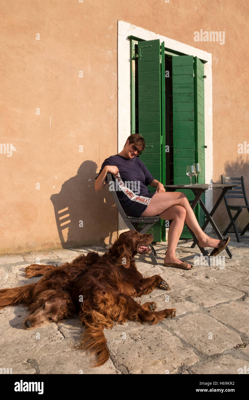 A woman enjoys a glass of wine in the sunshine with her Irish Setter dogs in the Old Town of Korcula, Croatia. Stock Photo