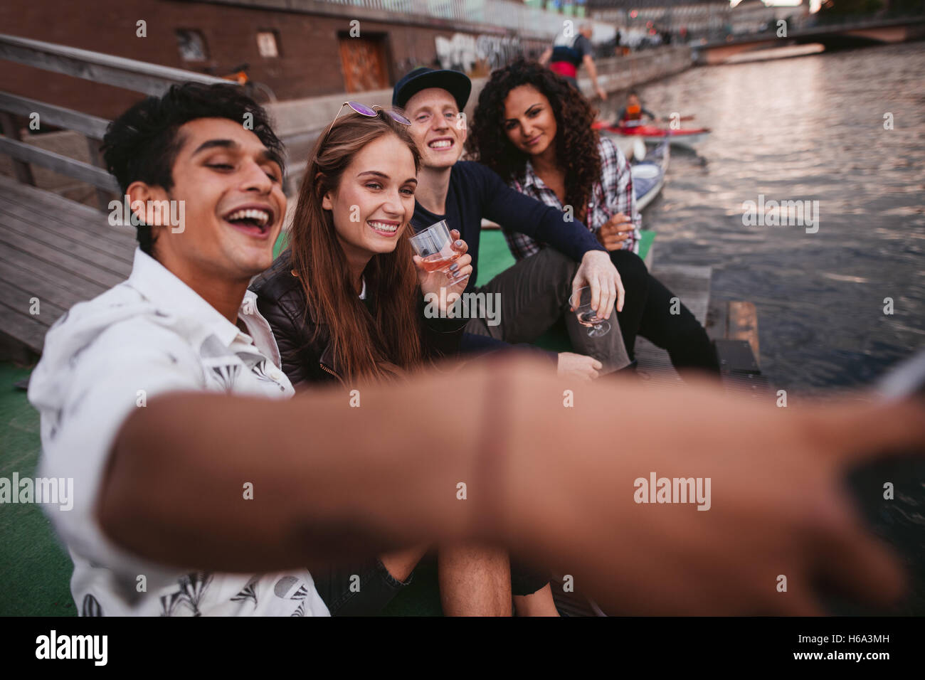 Shot of young man taking selfie with friends sitting on jetty on the lake. Teenagers having fun outdoors taking self portrait wi Stock Photo