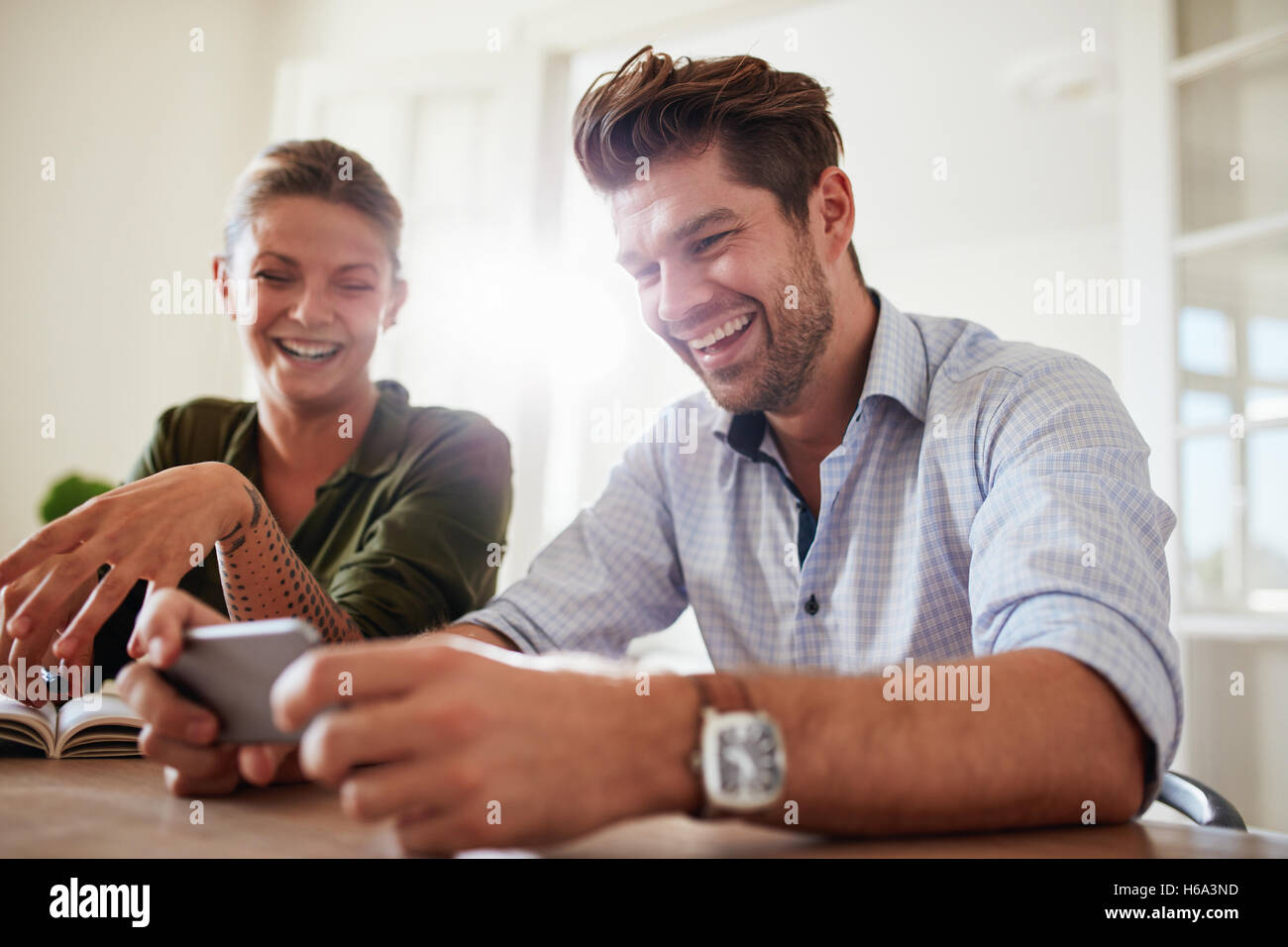 Shot of young couple sitting at table using cellphone at home and smiling. Man and woman smiling with mobile phone. Stock Photo