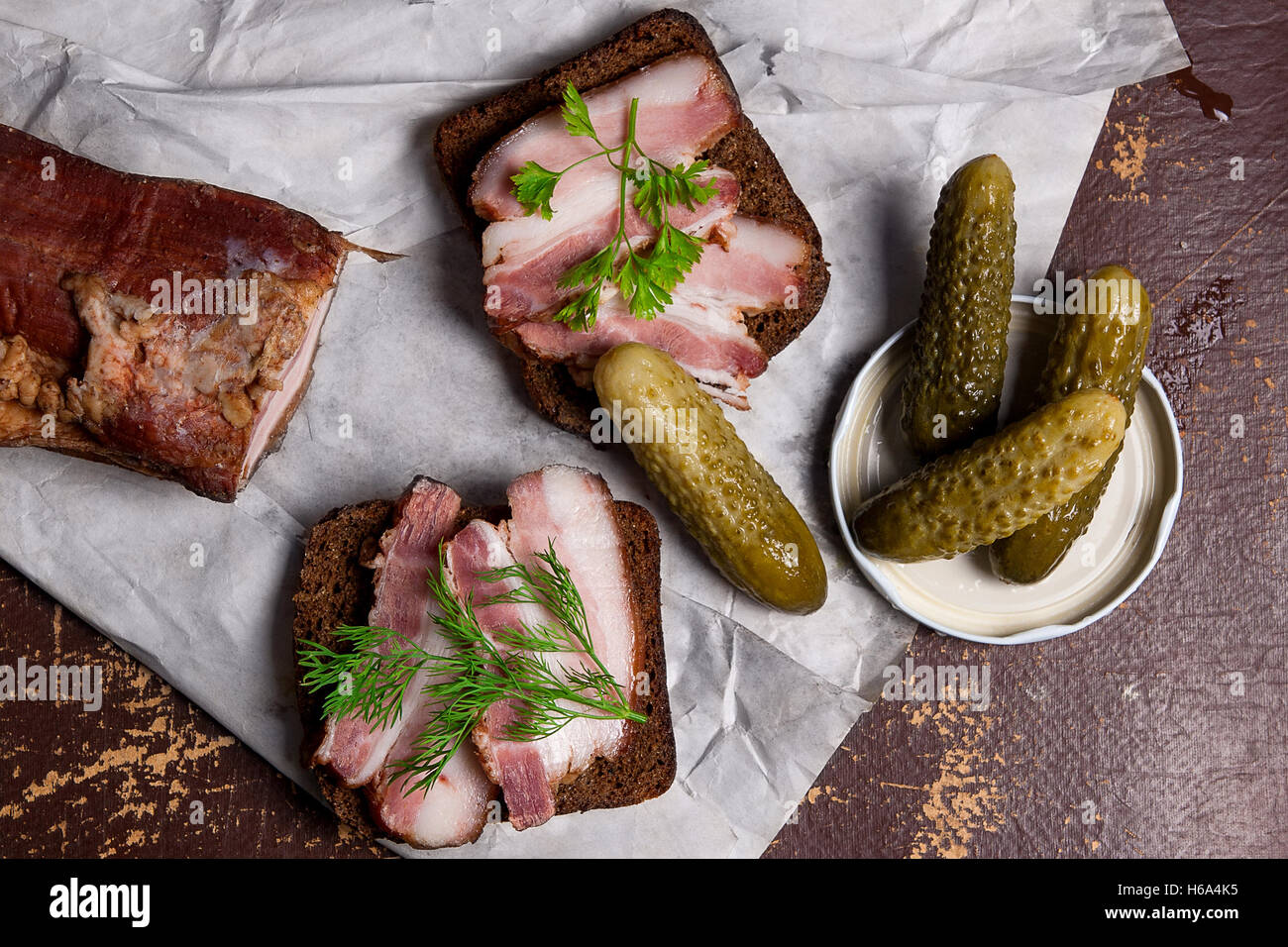 Close up view slices of smoked bacon on the piece rye bread with herbs and several pickles - rustic style Stock Photo