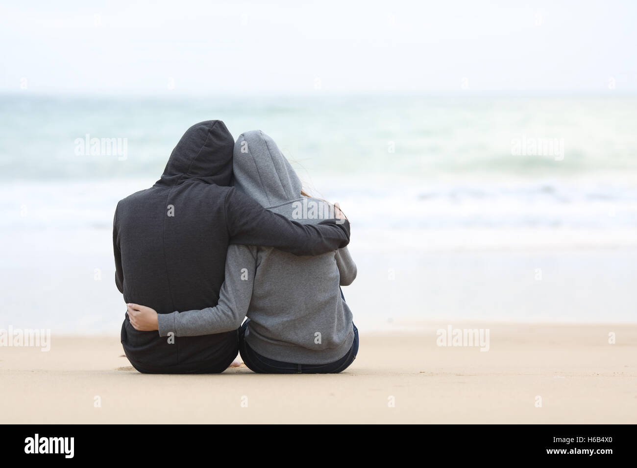 Rear view of a couple of pensive teenagers hugging and watching the sea sitting on the sand of the beach in a rainy day Stock Photo