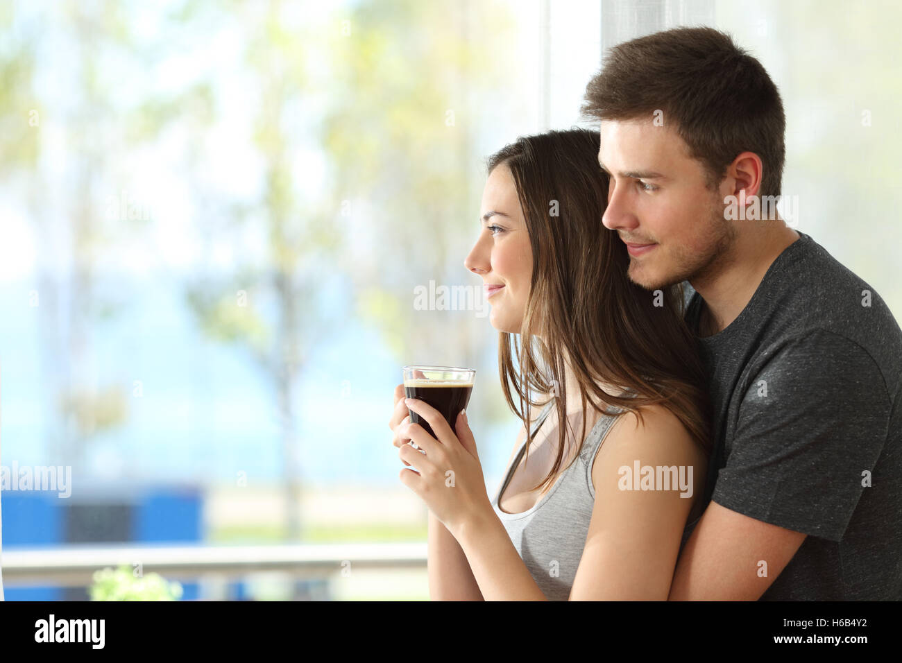Side view portrait of a pensive couple or marriage hugging and looking outdoors through a window of an hotel room Stock Photo
