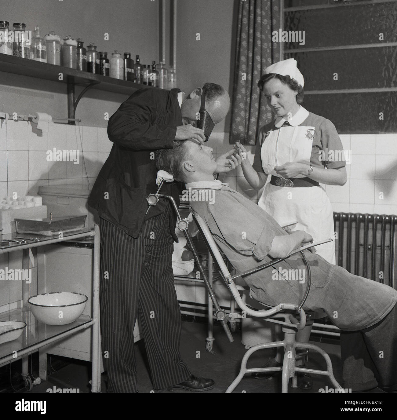 1950s, historical, a male factory worker in his overalls sitting on a chair in the company's surgery or medical room having his eyes checked by a male doctor, with a uniformed nursing sister in support, England, UK. Stock Photo