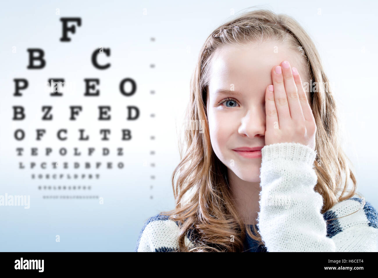 Close up portrait of girl reviewing eyesight closing eye with hand.Out of focus test chart in background. Stock Photo