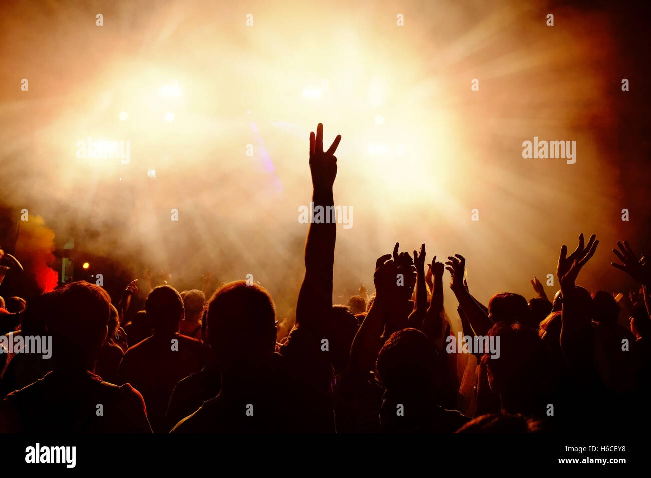 Silhouettes of festive crowd of young people dancing at music festival. Raising hands against smoky orange background. Stock Photo