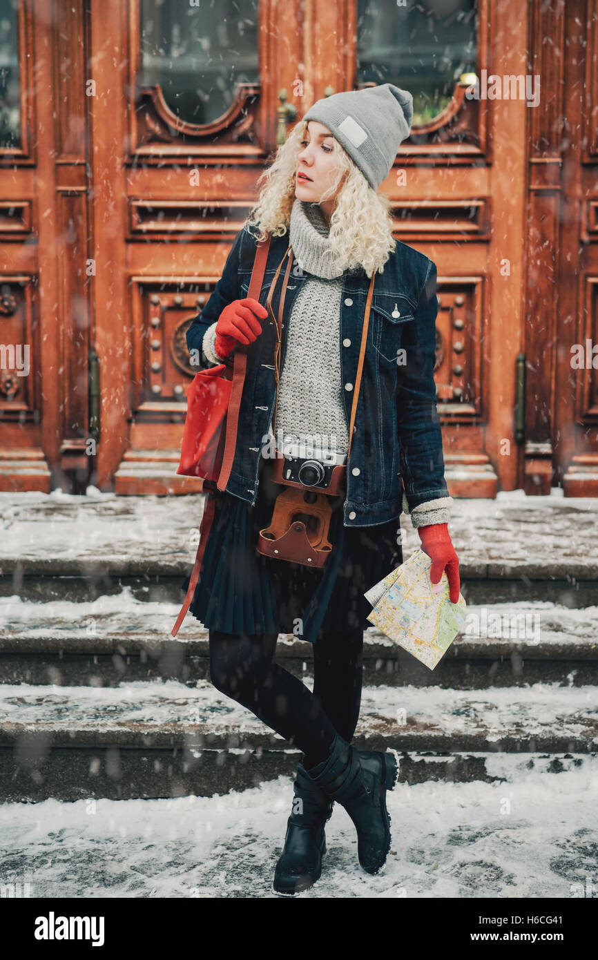 Young blond curly female in warm clothes with old film camera and map standing. Wooden doors with handles on the background Stock Photo