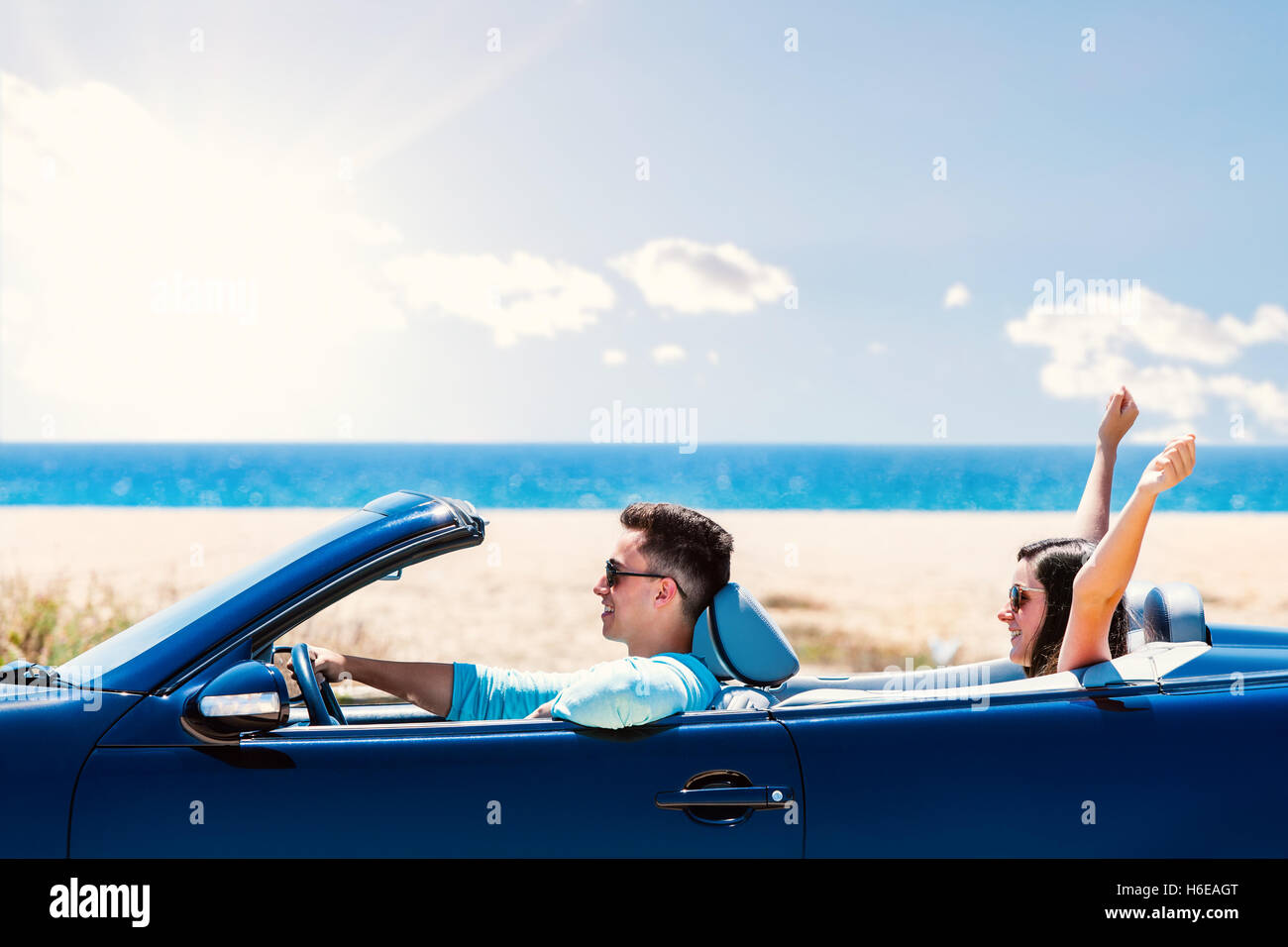 Close up portrait of young man driving blue convertible. Girlfriend sitting in back raising arms in air. Stock Photo