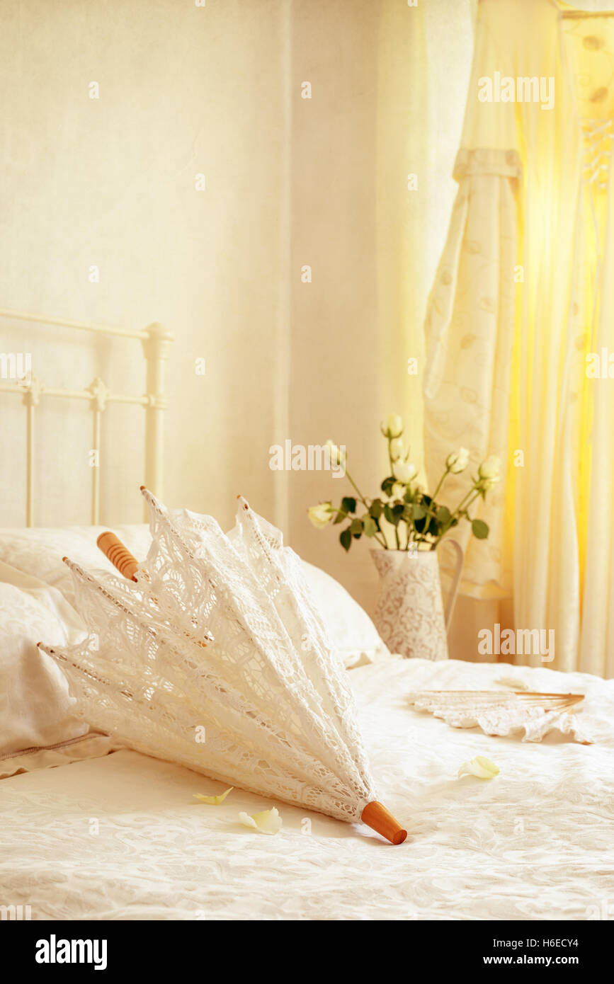 Brides parasol and fan lying on the bed with wedding dress hanging in the background Stock Photo
