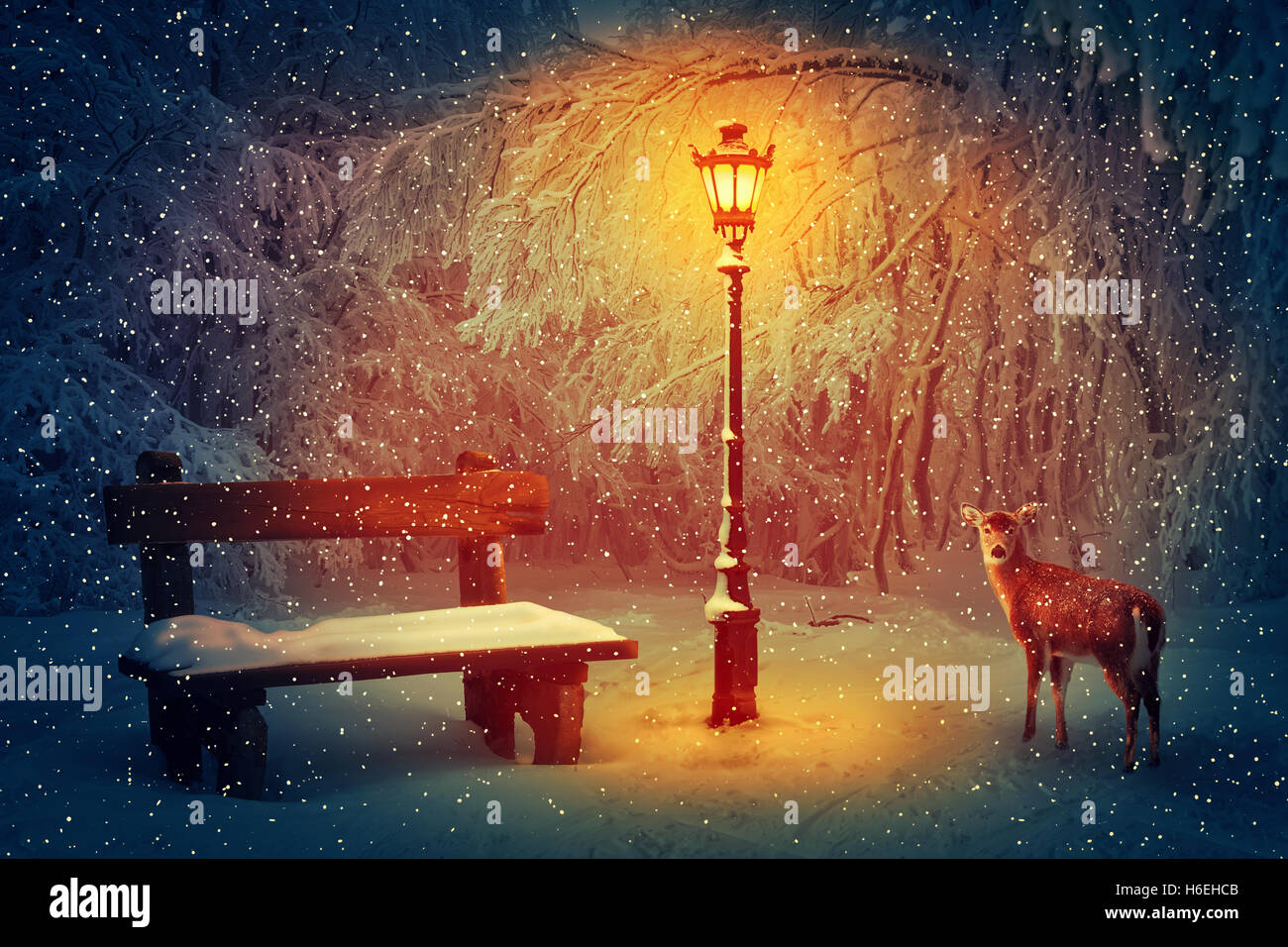 Wooden bench and a glowing lamp in the winter park covered in with snow. Snowing peaceful night scene and a doe looking carefull Stock Photo