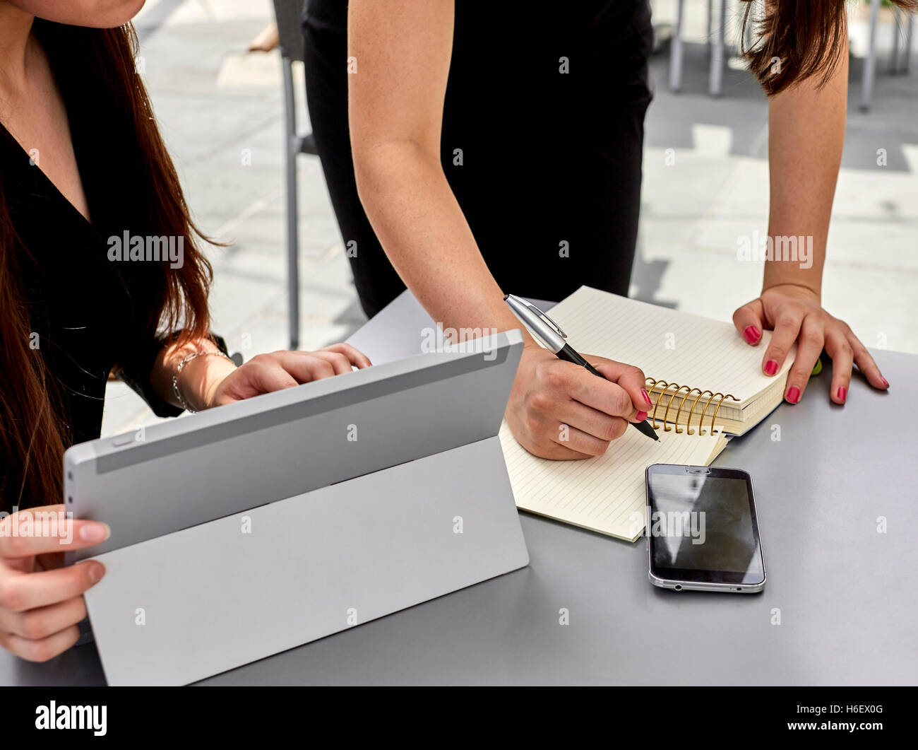 Two women one sitting one standing over a table prepared to write on paper while the other uses a tablet and a cell phone on the Stock Photo