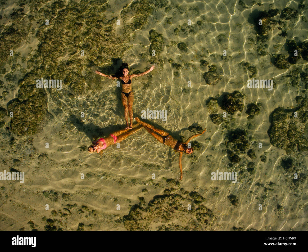 Aerial view of three young women lying in sea water making a formation. Female friends relaxing in sea with their legs together. Stock Photo