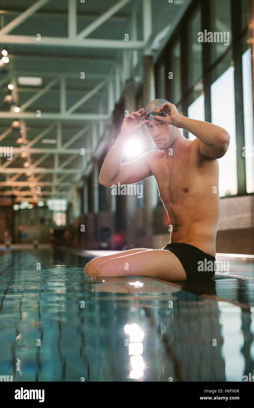 Shot of professional male swimmer sitting on edge of swimming pool, putting on swimming glasses. Stock Photo