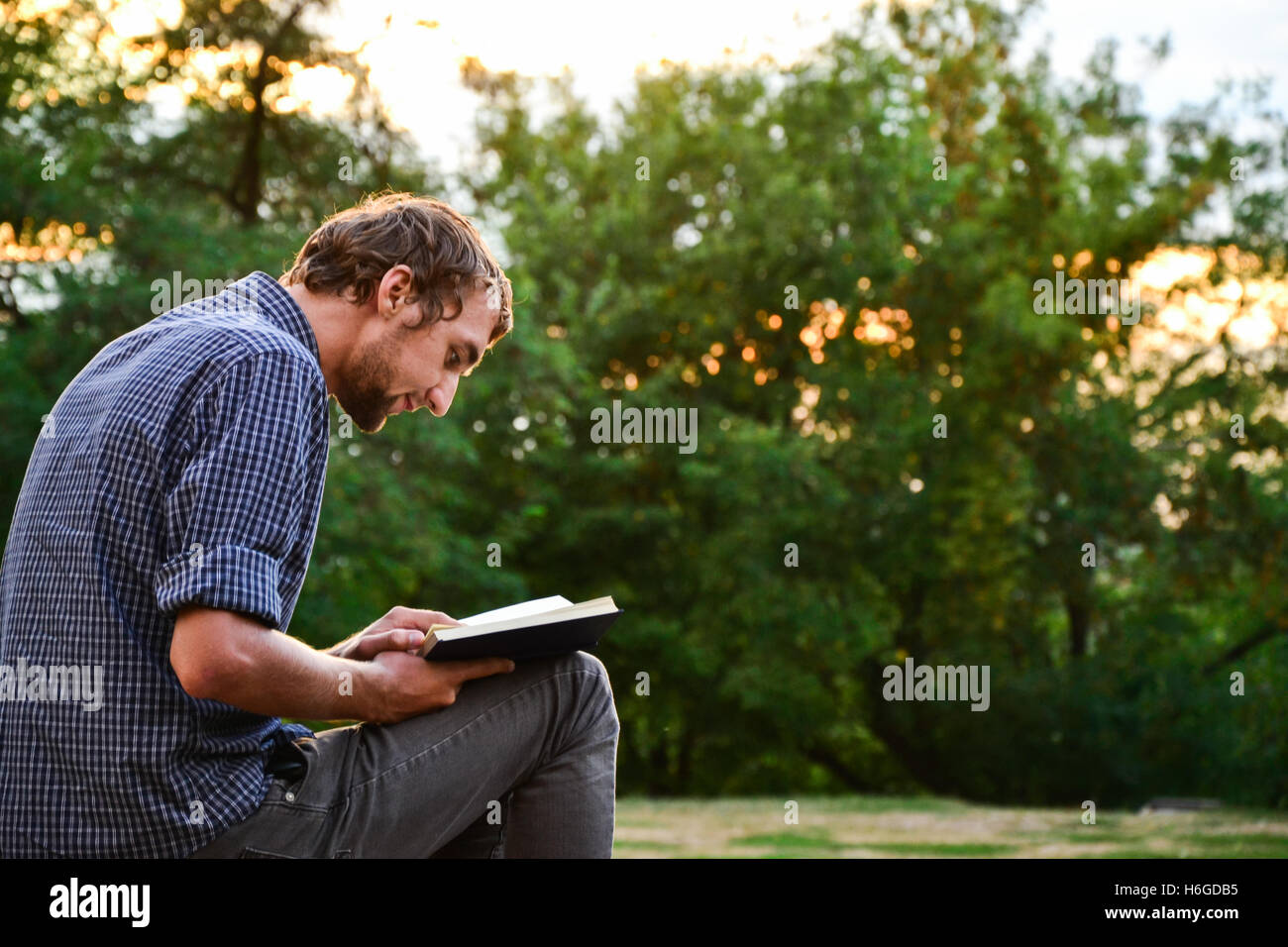 Guy sitting on a bench in the park reading book Stock Photo - Alamy