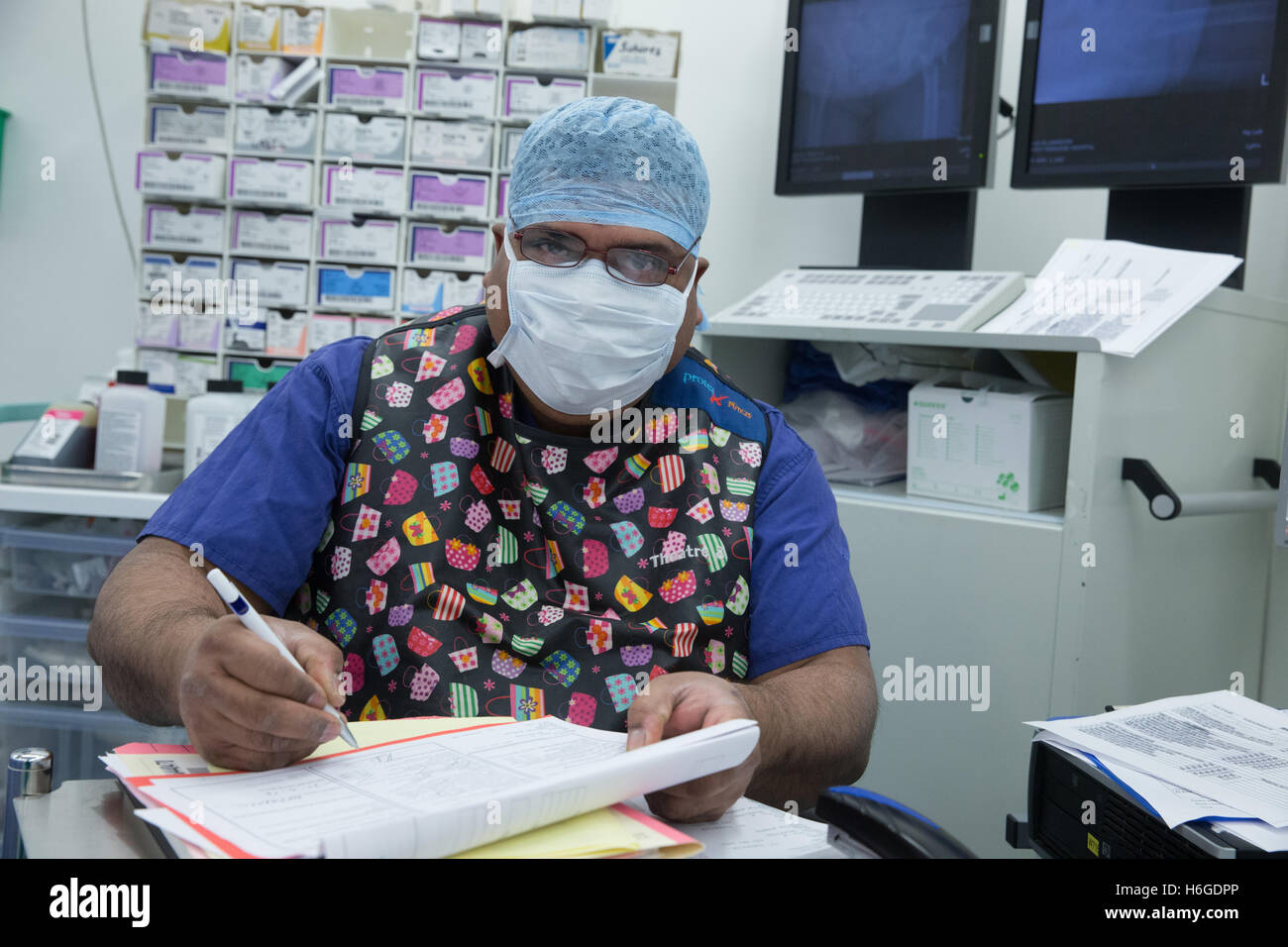 A hospital nurse wearing scrubs and a surgical mask in an operating theatre makes notes during an operation Stock Photo