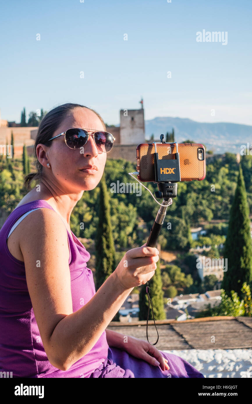 Female wearing sunglasses while sitting on the Mirador San Nicolás, taking selfie, Alhambra, Granada, Spain. Stock Photo