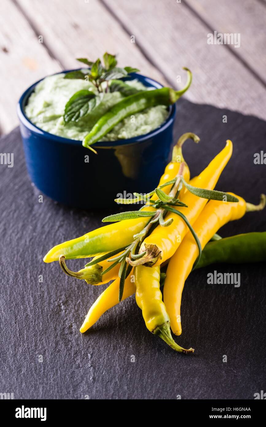 Vertical photo of wasabi dip in small blue bowl. Green chili pepper is placed on dip with green mint herb. Bunch of yellow peppe Stock Photo