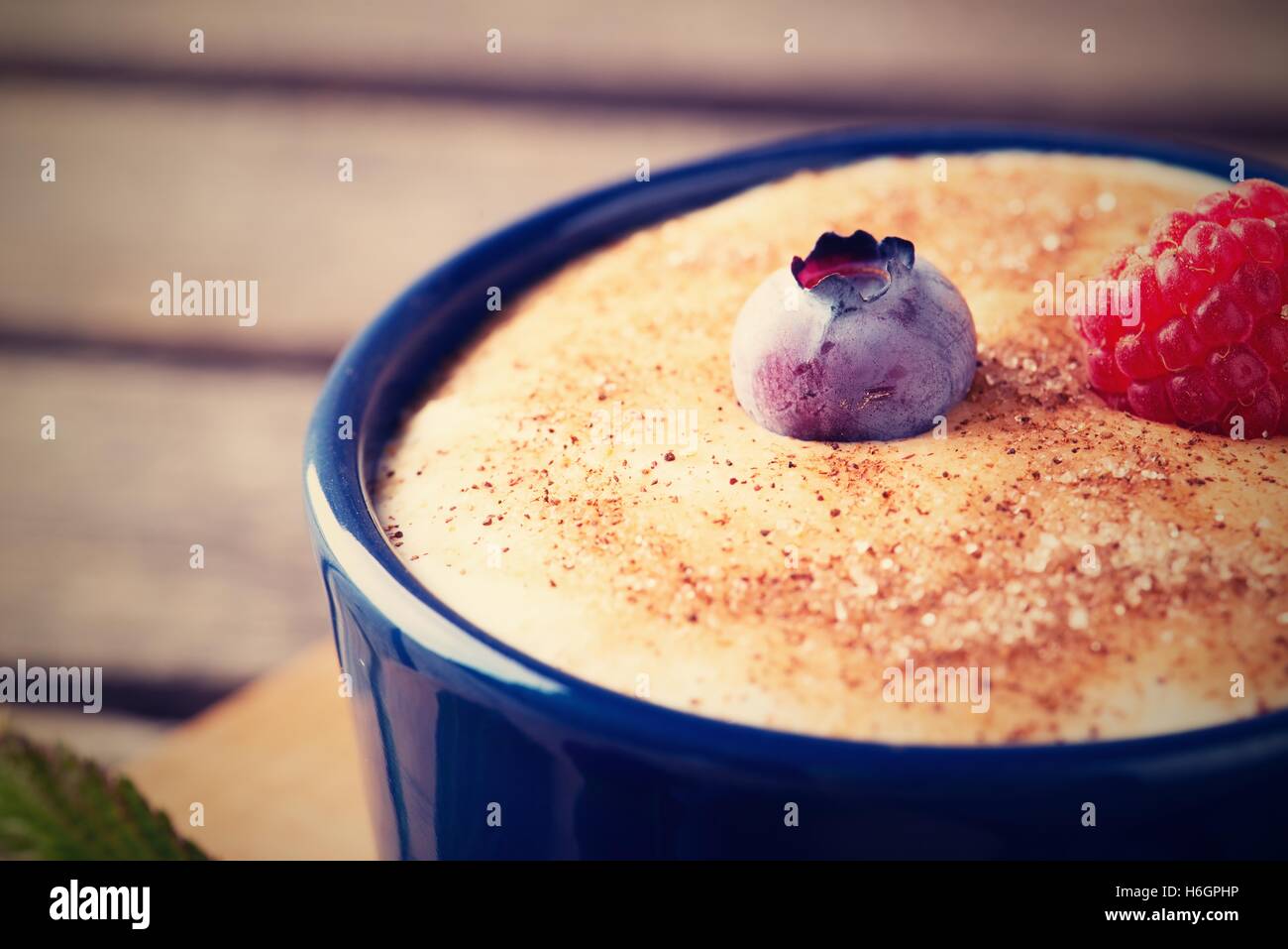 Horizontal vintage retro photo of creamy dessert in blue bowl. Cinnamon sugar is on the surface with single blueberry and red ra Stock Photo