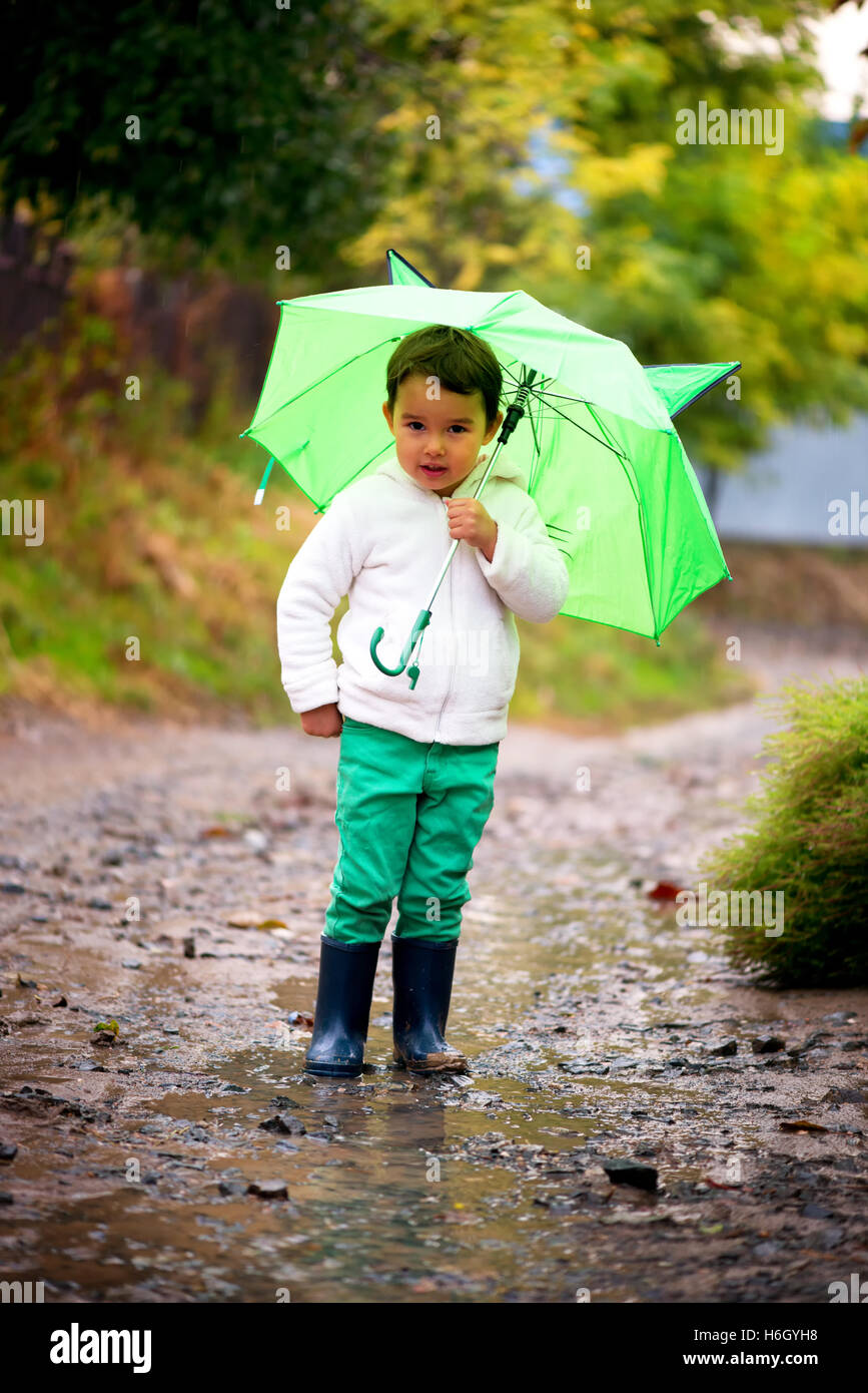 baby girl with an umbrella in the rain runs through the puddles playing on nature Stock Photo