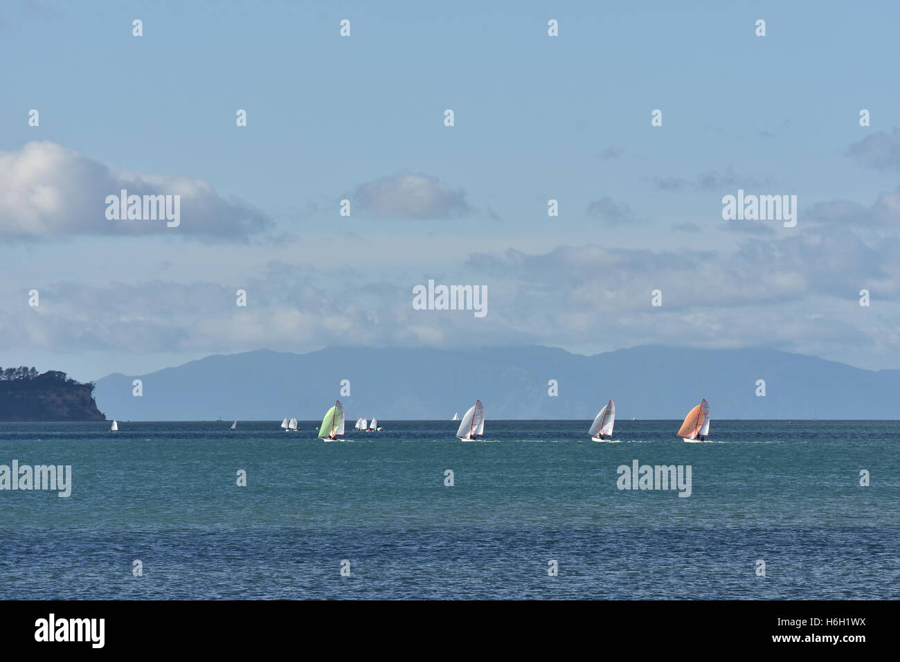 Sailboats with sails of various colours sailing on calm blue-green sea with big landmass in background. Stock Photo