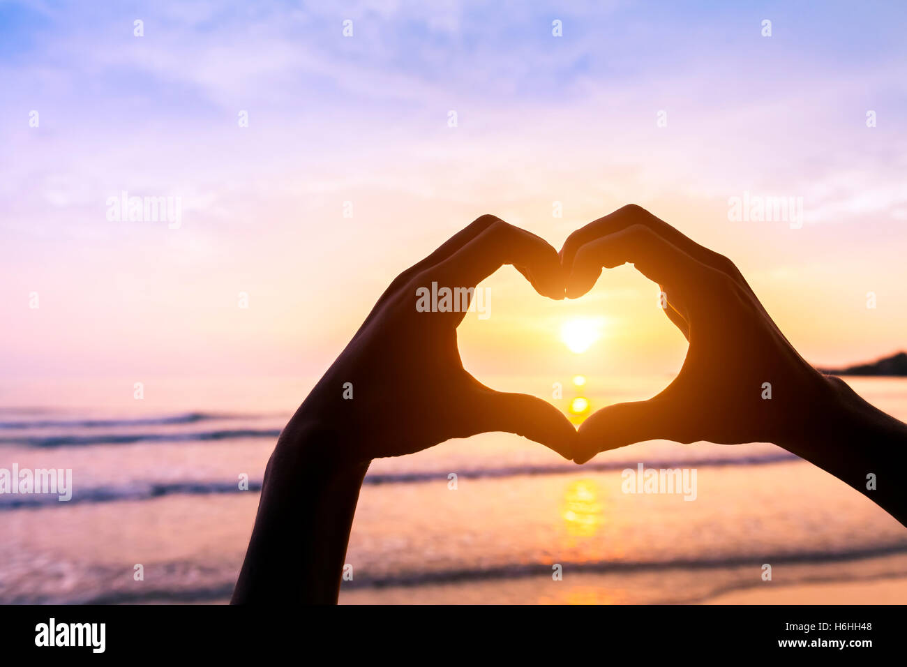 Shape of heart done with silhouette of hands on a beach with sunset sun and colors - Symbol of love - Romantic travel Stock Photo
