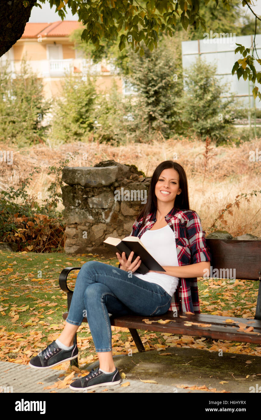 Beautiful young woman reading a book sitting on a bench in park at fall ...