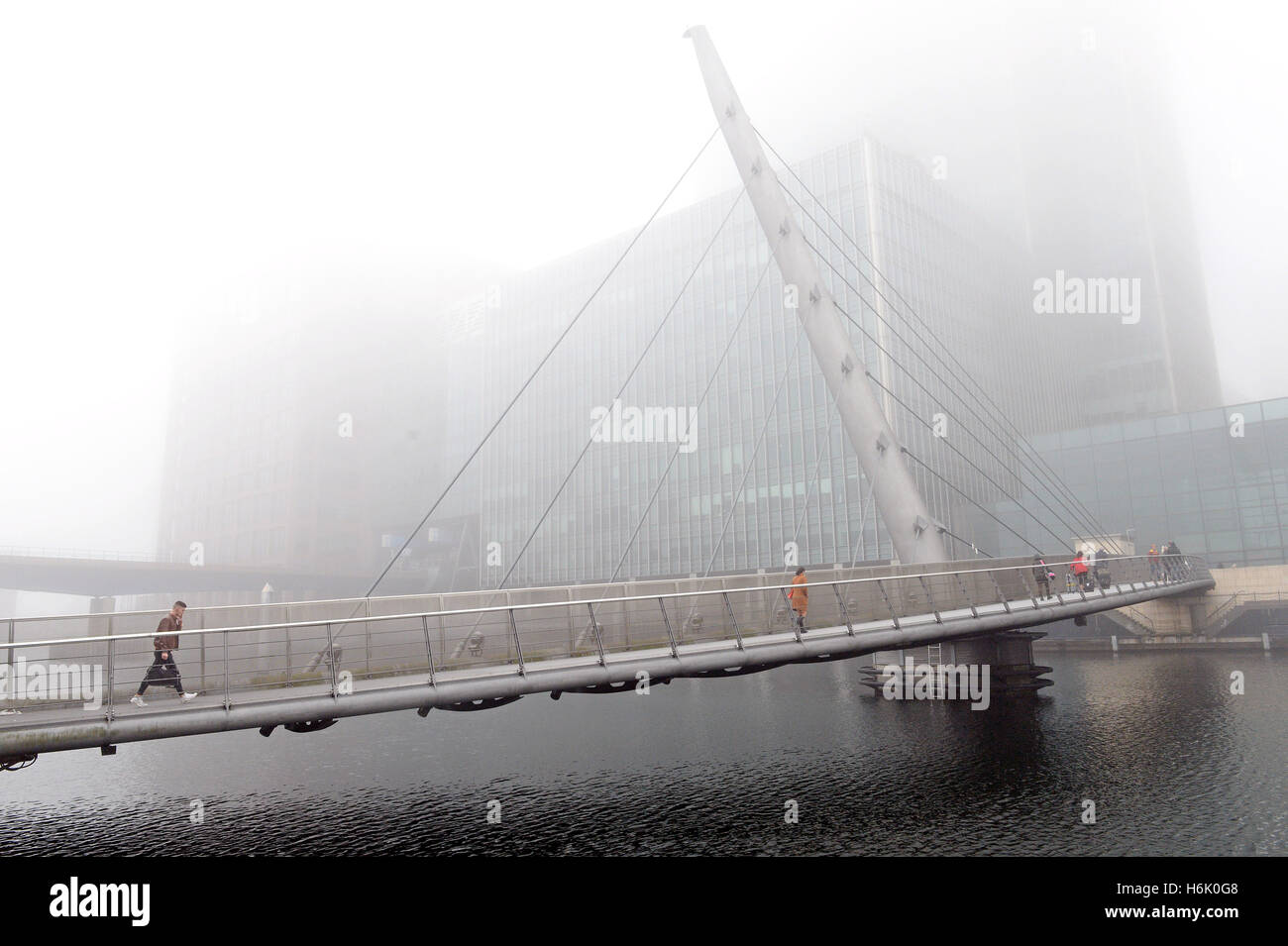 People walk across the swing bridge in Canary Wharf, London, as mist obscures the top of the buildings. Stock Photo