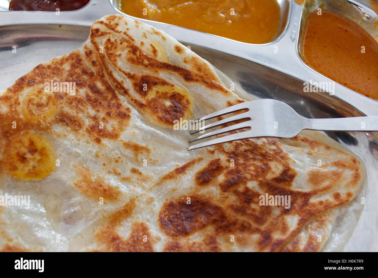 Roti canai or pan fried flatbread consisting of dough, egg, ghee and banana with the three accompanying sauces Stock Photo