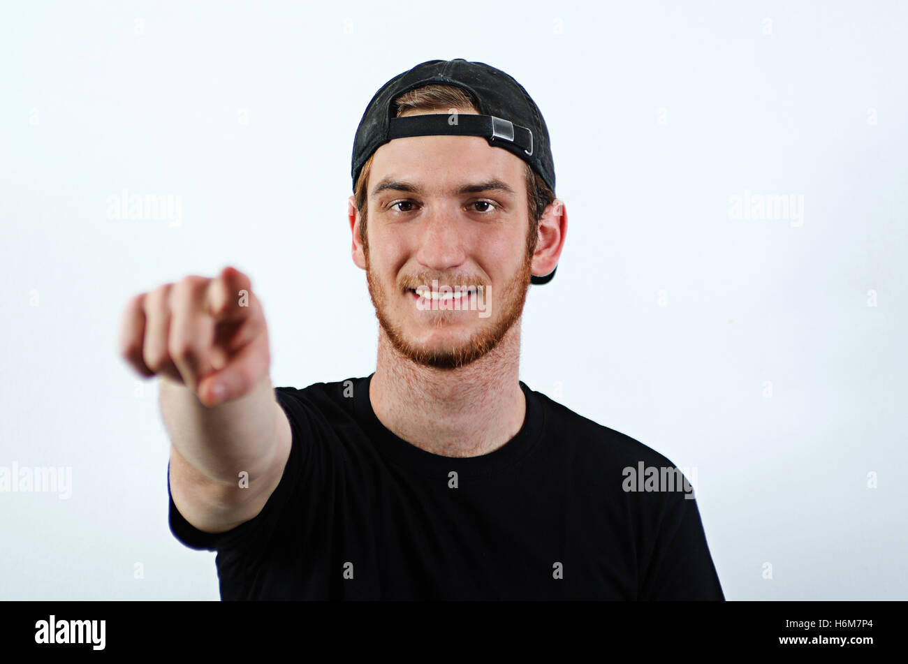Smiling Young Adult Male in Dark T-Shirt and Baseball Hat Worn Backwards Pointing Towards the Viewer Stock Photo