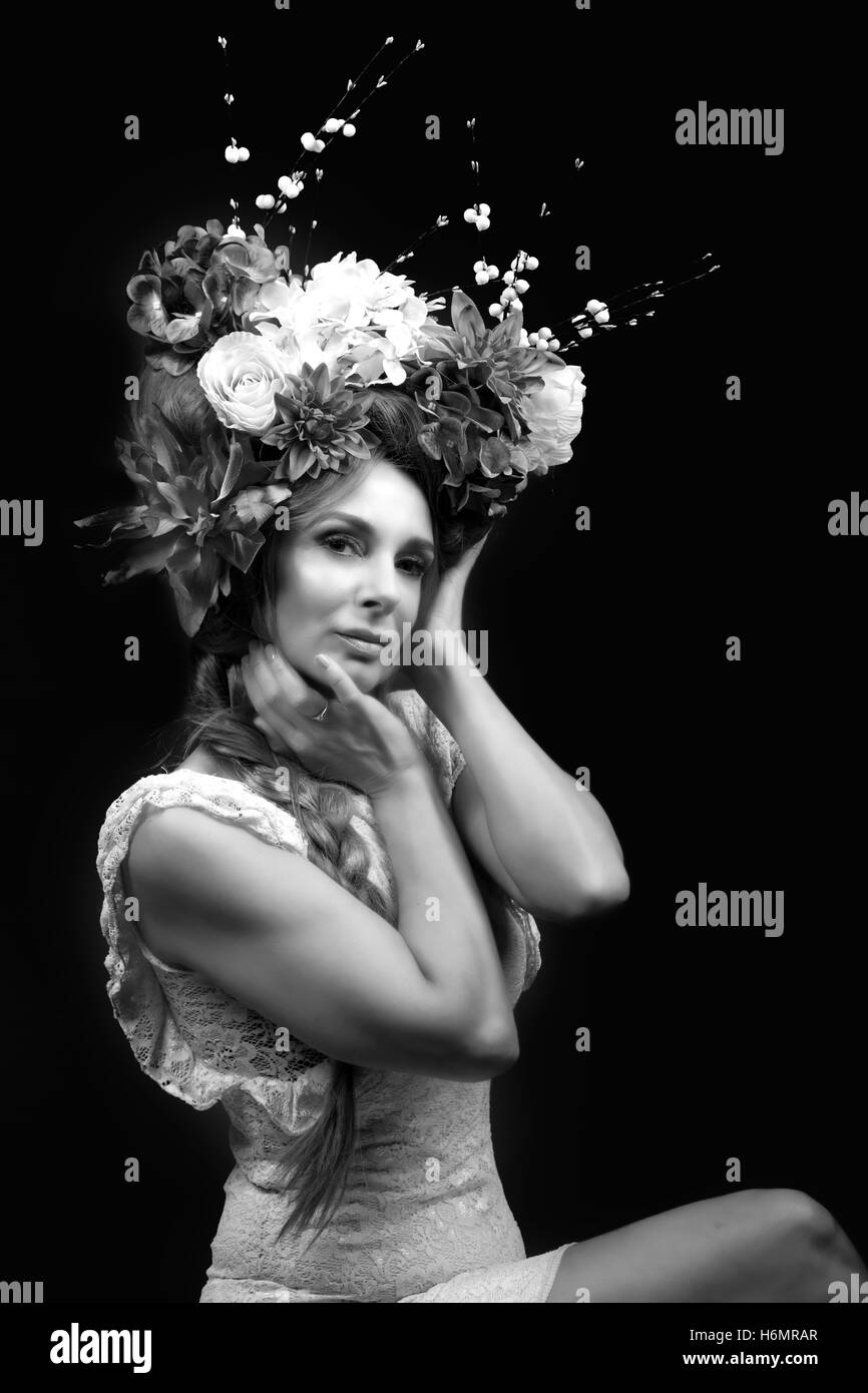 Model woman with architectural floral hair piece wearing white lace dress photoshoot against black studio backdrop, monochrome Stock Photo