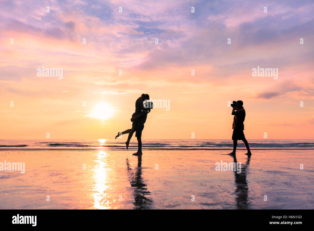 Photographer taking photo of a couple kissing on the beach during sunset - concept about romantic travels and honeymoon Stock Photo