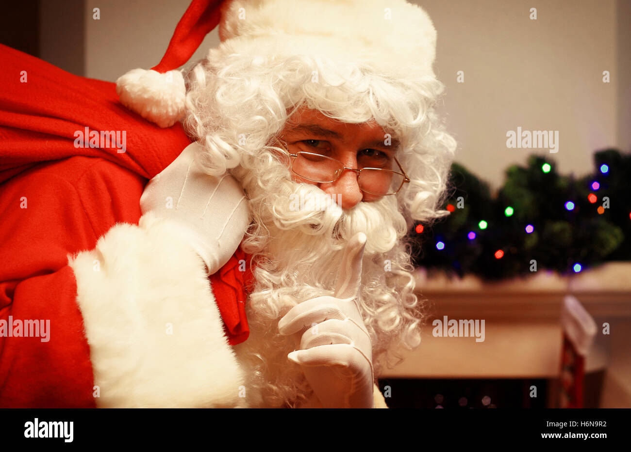 Portrait of Santa Claus with huge red sack keeping forefinger by his mouth and looking at camera. Stock Photo