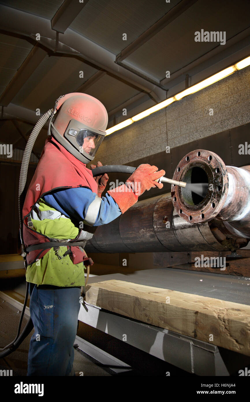 A worker uses high-pressure grit-blasting to clean a section of refinery pipe at a petrochemical maintenance facility Stock Photo