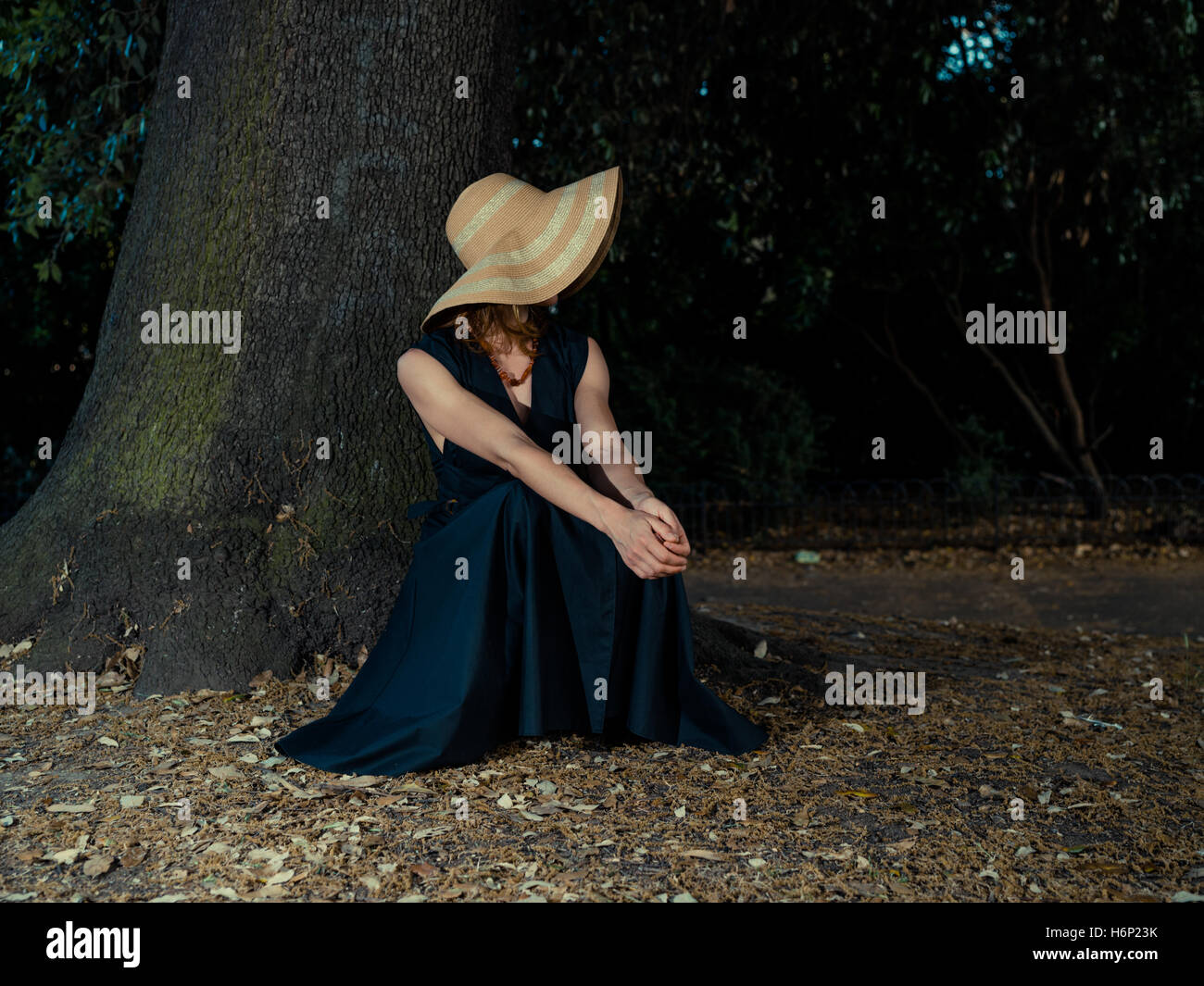An elegant woman wearing a dress and a hat is relaxing under a tree in ...