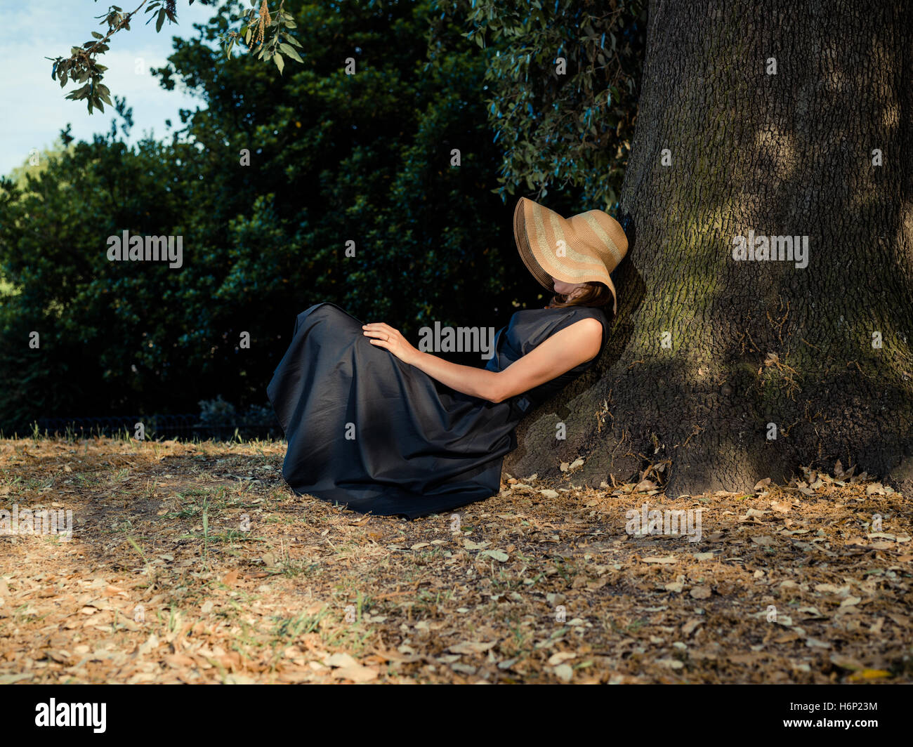 An elegant woman wearing a dress and a hat is relaxing under a tree in ...
