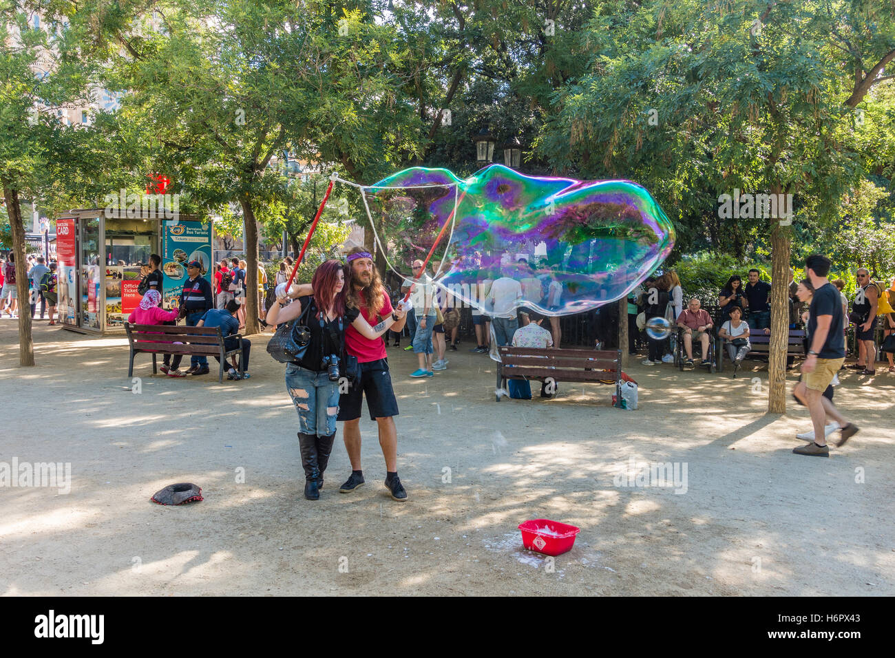A young twenty-something Spanish woman is helped by a man in making enormous soap bubbles in a park by the Sagrada Familia, Stock Photo