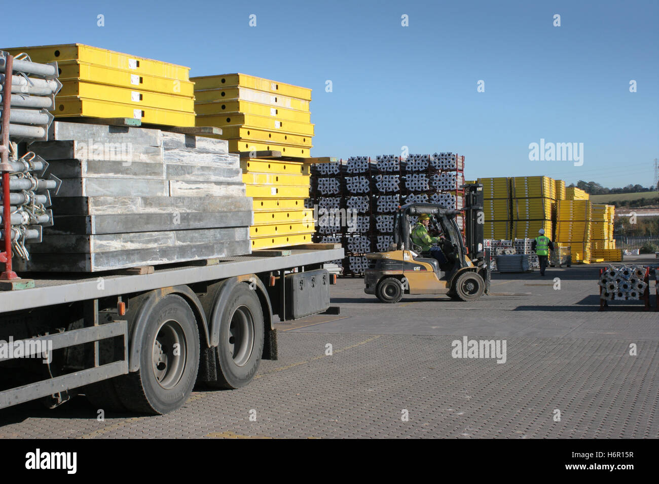 A forklift truck unloads steel formwork products from a flatbed lorry in a UK storage yard. Bright, sunny day. Stock Photo