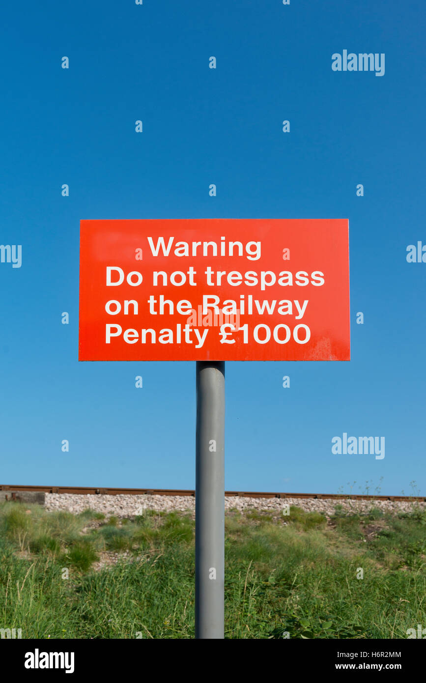 Red Warning Do Not Trespass on the Railway Penalty £1000 sign on pole with railway line in the background against clear blue sky Stock Photo