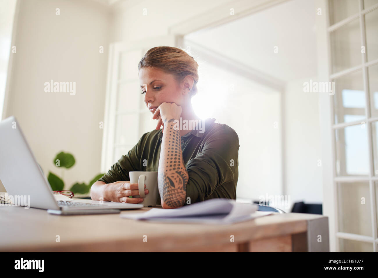 Indoor shot of young woman with coffee looking at laptop. Female sitting at table working. Stock Photo