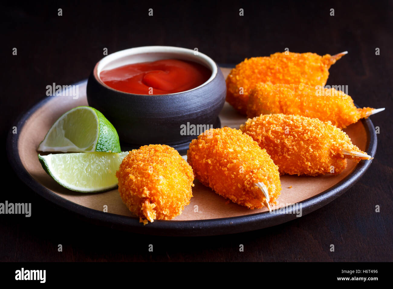 Rustic plate of fried breaded surimi crab claws with bowl of tomato sauce and lemon slices isolated on black. Stock Photo